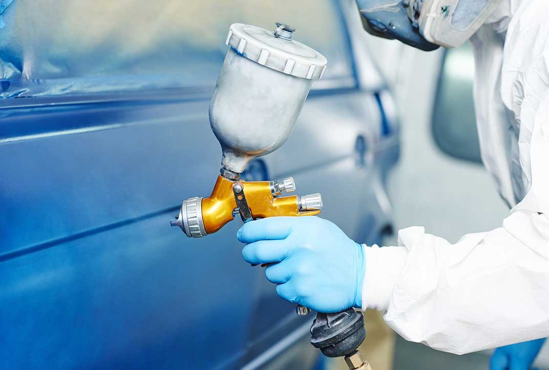 A person in protective clothing applying paint to a car. Only the person’s hand and arm are shown.