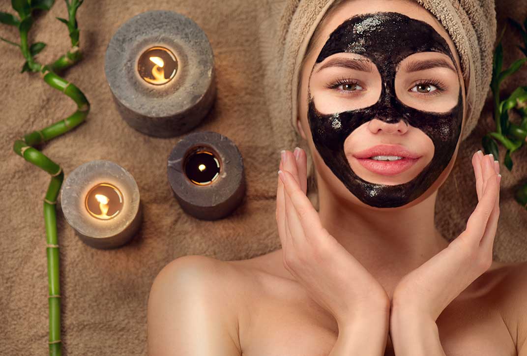 A woman with a dark face mask lies on a bath towel. A bath towel is wrapped around her head. Next to her are three candles.