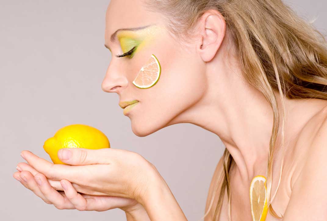 A woman with yellow made-up lips and eyelids and a slice of lemon on her cheek smells a lemon she holds in her hands.