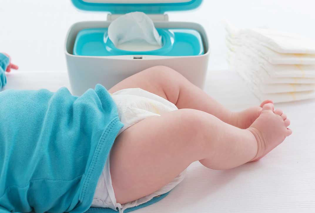 Der Unterkörper eines Babys auf einem Wickeltisch. Im Hintergrund steht ein Spender mit Feuchttüchern, daneben ist ein Stapel Windeln.