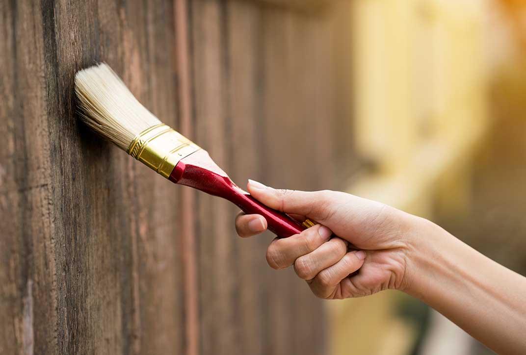 The photo shows a hand painting a fence with a paintbrush.
