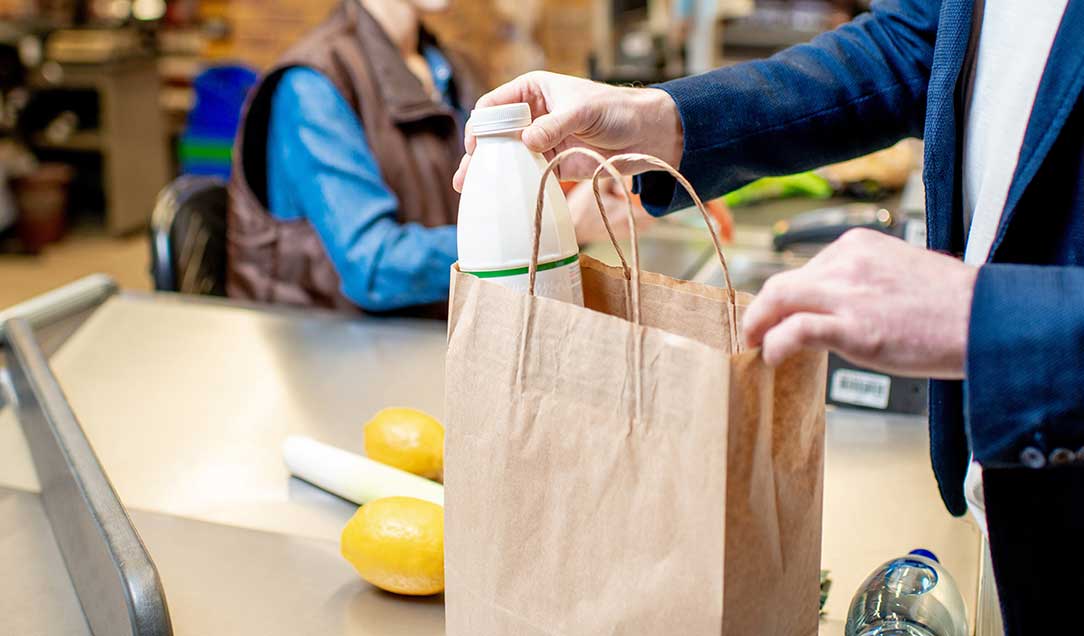 Eine Supermarktkasse. Im Hintergrund sieht man eine Kassiererin, vorne packt ein Mann eine Milchflasche in eine Papiertüte.