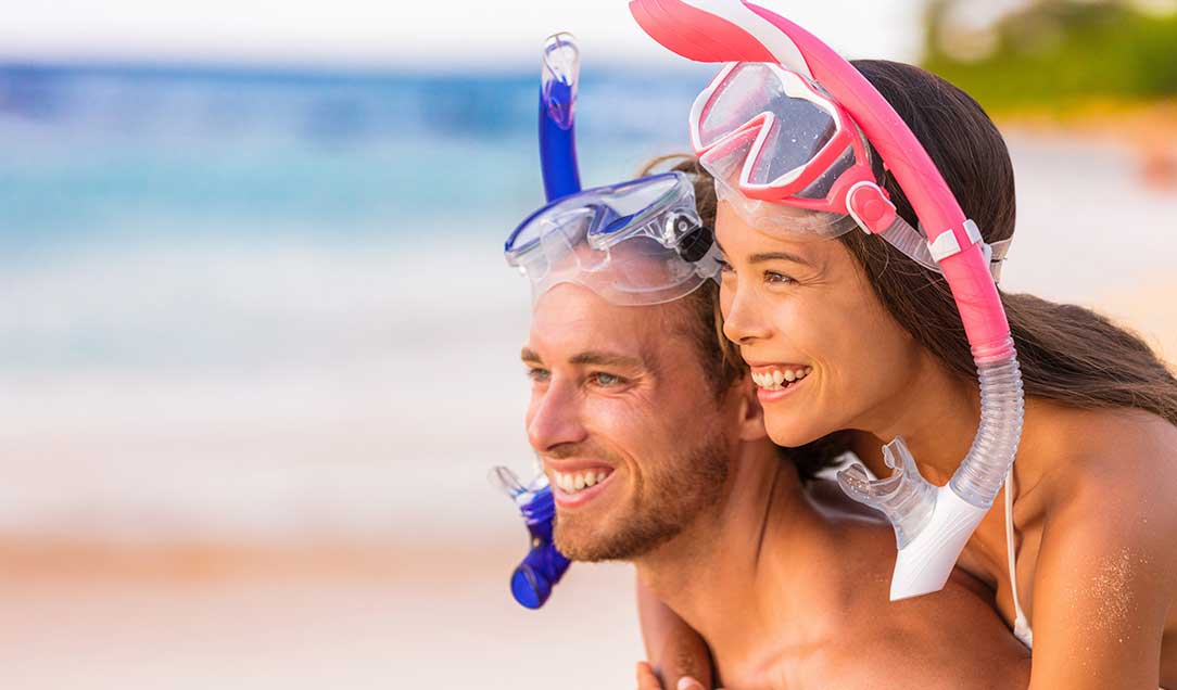 Eine Frau und ein Mann mit Taucherbrillen und Schnorcheln am Strand. Sie schauen in die Ferne und lächeln.