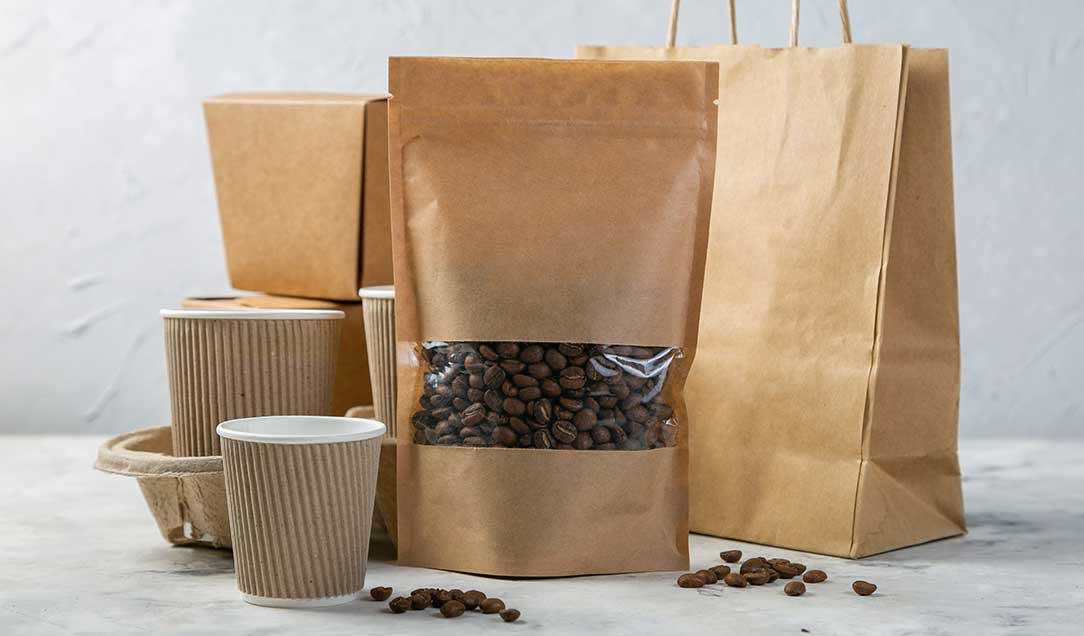 Various paper packaging: bags, folding boxes, pouches and cups. A stand-up pouch contains coffee beans that can be seen through a transparent window. Some coffee beans lie in front of the packaging.