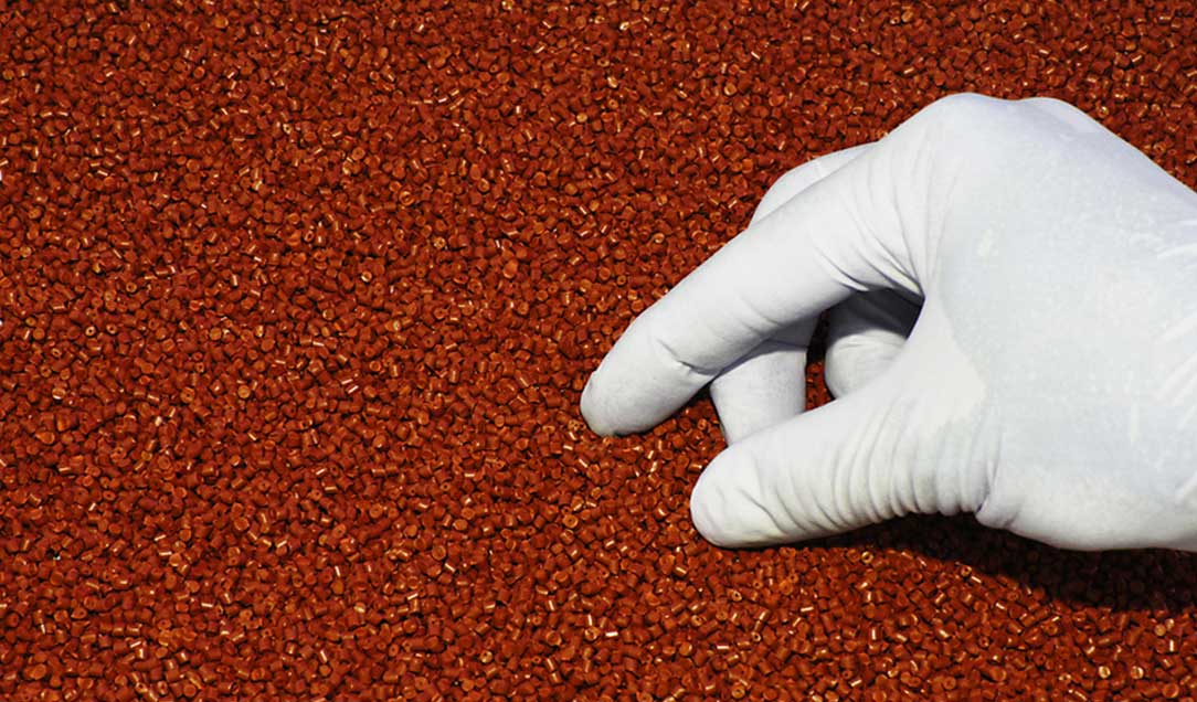 Eine Hand mit weißem Handschuh greift in rotbraunes Kunststoff-Granulat