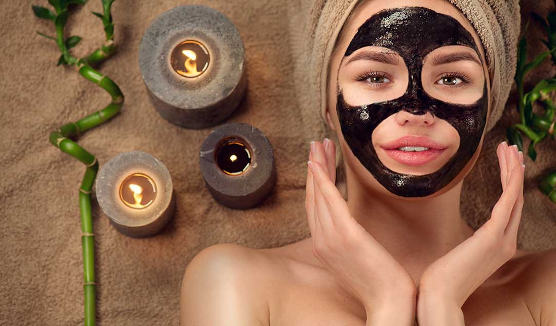 Frau mit einer dunklen Gesichtsmaske und einem Badetuch um den Kopf gewickelt liegt auf einem Badetuch. Neben ihr stehen drei Kerzen.
