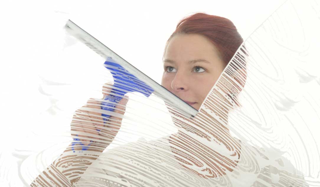 Eine junge Frau steht hinter einer eingeseiften Glasscheibe und beginnt sie mit einem Abzieher zu reinigen.