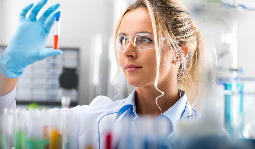 Frau in Laborkittel, mit Handschuhen und Schutzbrille hält ein Reagenzglas hoch, das mit einer roten Flüssigkeit gefüllt ist, und betrachtet es.